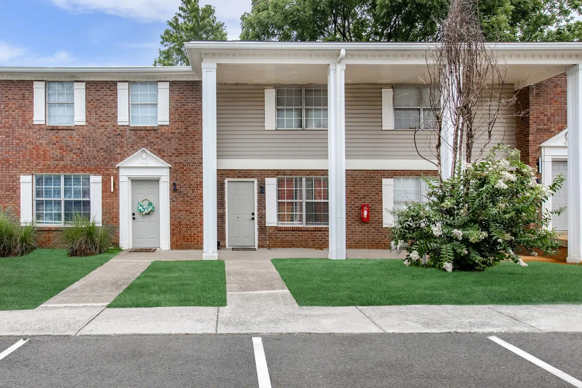 A warm exterior shot of a brick facade with white columns and a front door framed by lush green landscaping. The inviting front porch offers space for socializing or a quiet coffee, and the surrounding garden sets a tranquil, residential tone.