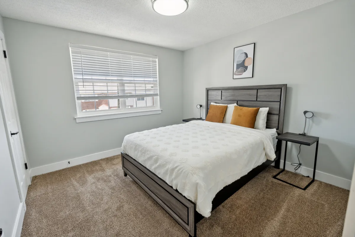 The interior shows a serene bedroom with a neatly made bed, a cozy nightstand, sunlight streaming through a large window, and tasteful wall art, creating an inviting, tranquil atmosphere ideal for rest and relaxation.
