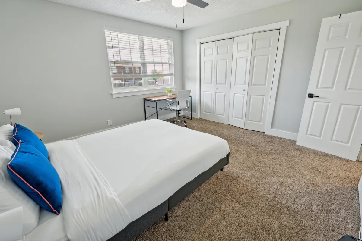 A sunlit bedroom adorned with blue accent pillows, a neatly made bed, a desk with a plant for a touch of freshness, and large closet doors for ample storage. The overall space is organized, serene, and ideal for both rest and productivity.