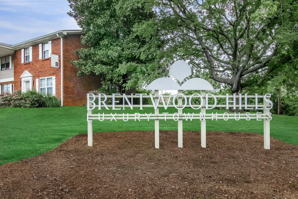 A welcoming entrance to Brentwood Hills, featuring a prominent sign surrounded by lush greenery and distinctive townhouse architecture in the background. The landscaping and communal areas invite residents to gather and socialize outdoors.