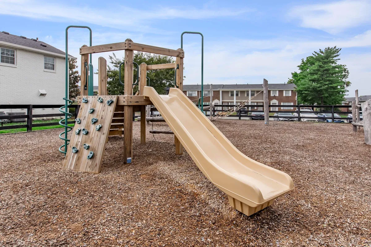 A vibrant playground structure with a slide and climbing wall sits amidst a leafy outdoor setting. The scene is lively, with space for children to play and families to gather. Benches and open areas invite group picnics or relaxing while kids explore.