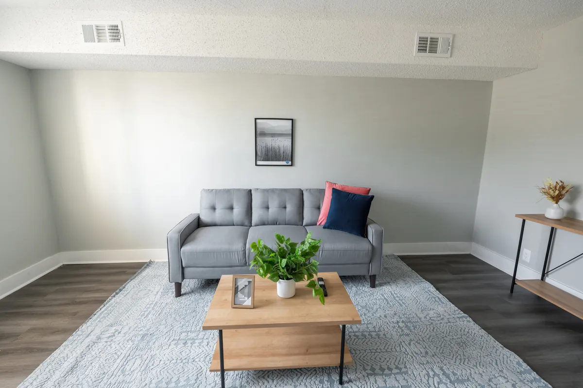 A bright, contemporary living room featuring a spacious grey couch, a wooden coffee table, lush green plant, and modern wall art. The room is filled with gentle natural light, creating an inviting atmosphere ideal for both socializing and quiet moments. The furnishings are stylish yet comfortable, perfectly suited for gatherings or relaxation.