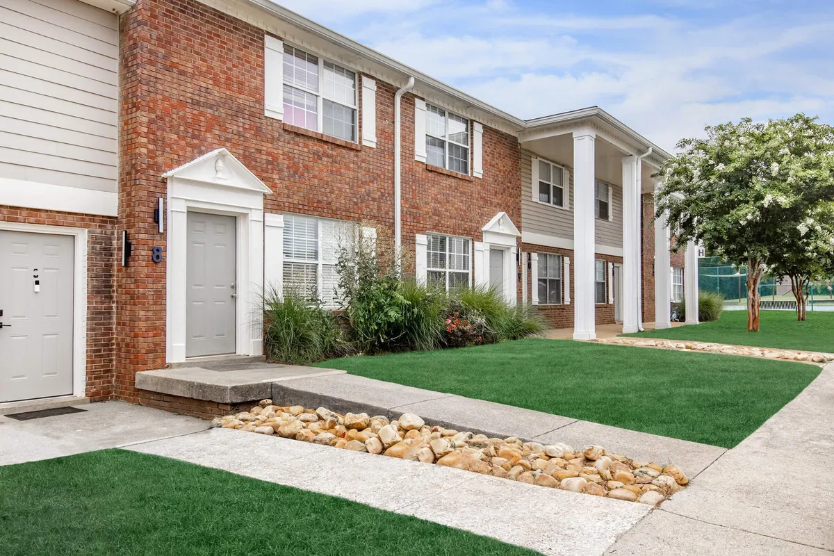 The image shows a welcoming exterior view of a brick townhouse with front doors and a landscaped garden. The scene is warm and inviting, featuring walking paths and greenery that suggest a peaceful, family-friendly community. This setting is ideal for casual socializing and sets the tone for relaxed gatherings.