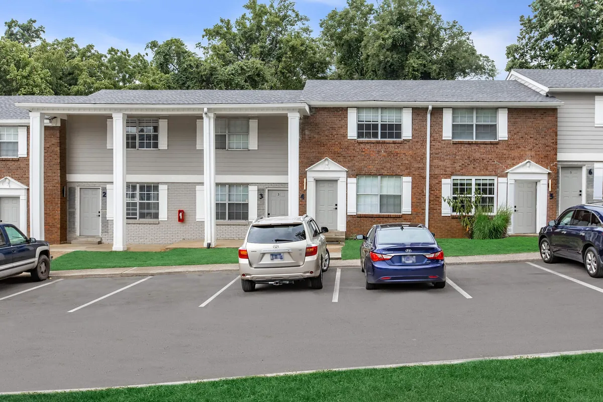 An exterior view of a two-story residential building with parked vehicles, green lawn, and a welcoming pathway leading to the front doors. The yard hints at community life, with space for socializing or moments of peace outside.
